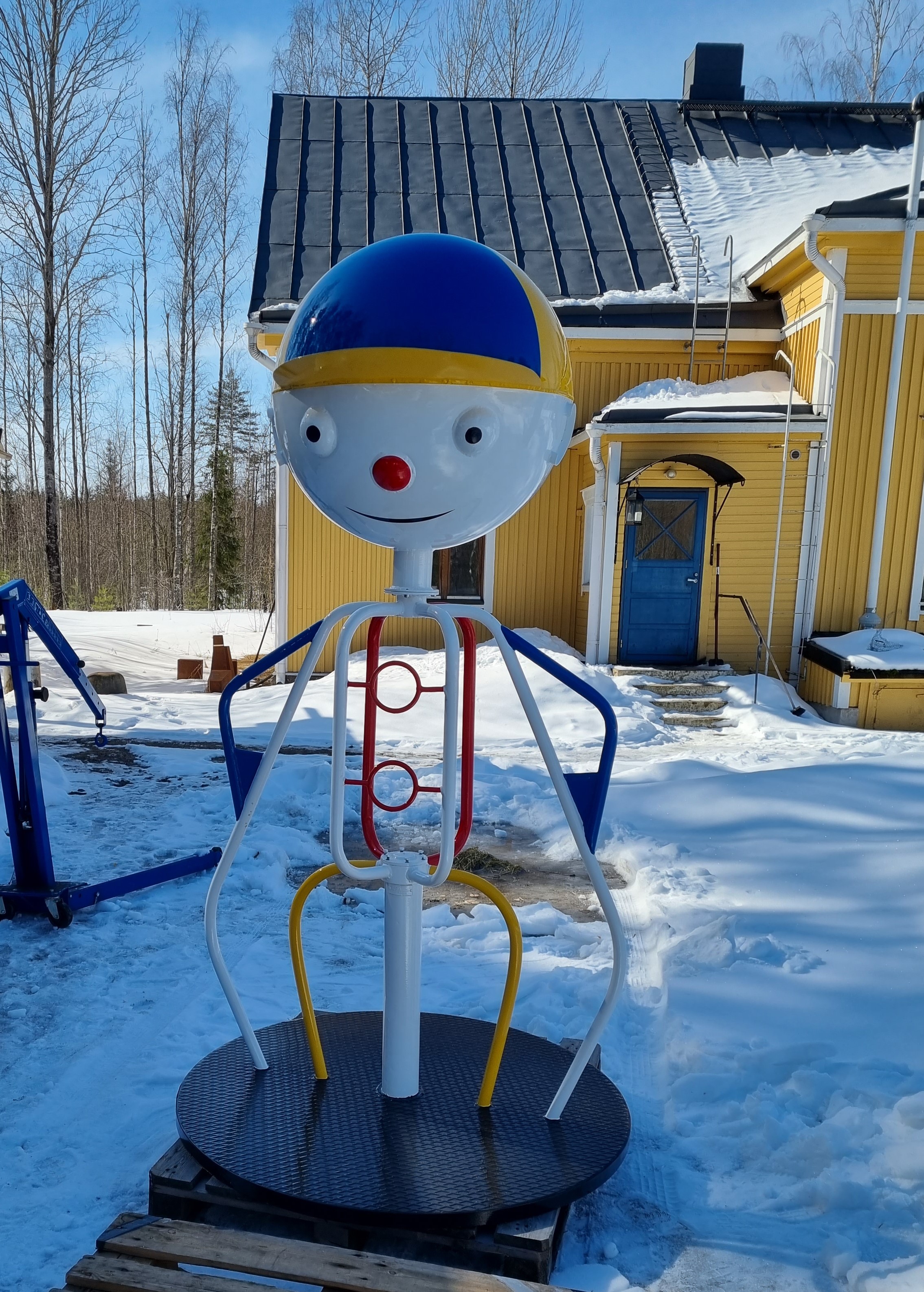 Metallinen, ihmismallinen lasten karuselli, jolla iso pää, silmät, nenä, suu ja lippahattu. Puistossa talvella, taustalla keltainen puutalo.
